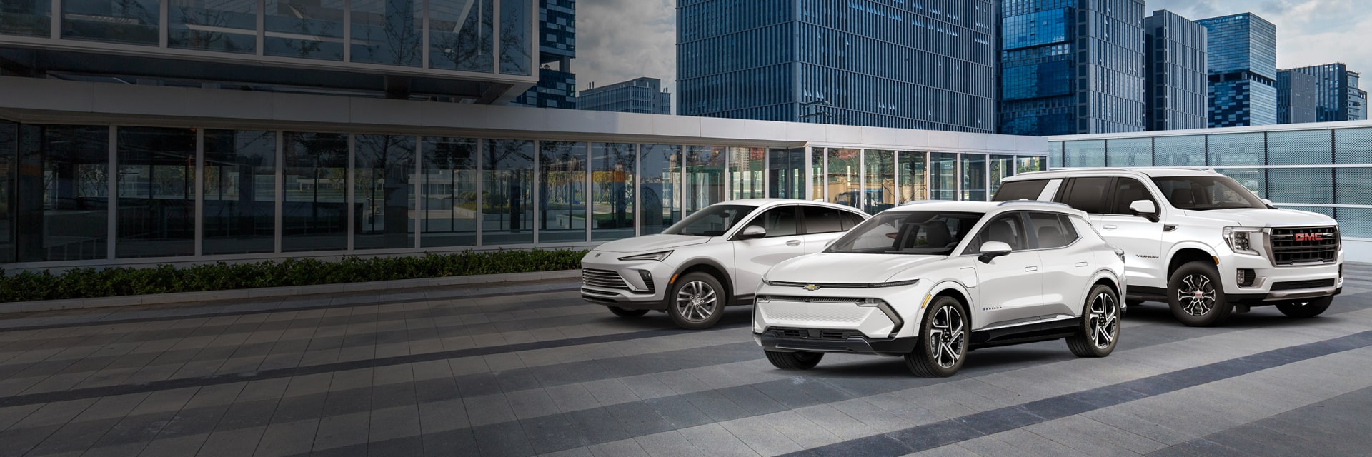Three white GM vehicles in front of city buildings